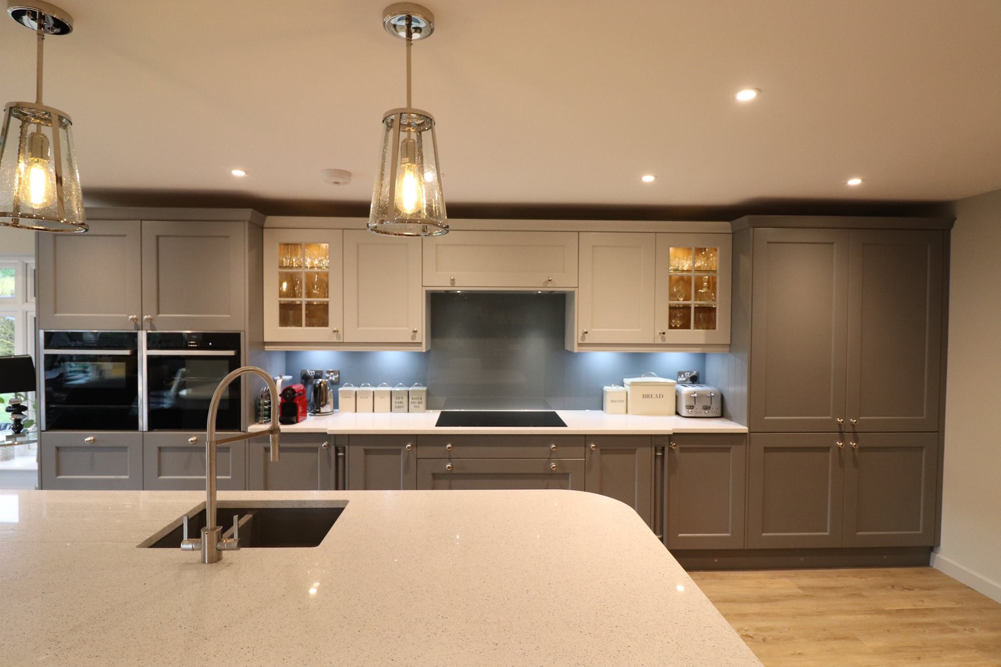 Warm grey painted in-frame shaker kitchen with glazed display cabinets, white quartz worktop and glass lantern pendants — Inner Peace Interiors, Camberley