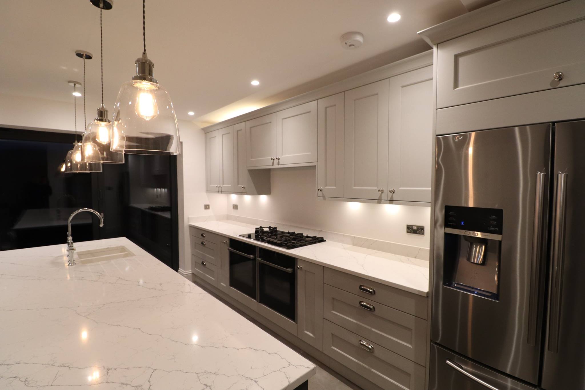 Light grey shaker kitchen with white quartz island worktop, glass dome pendant lights and integrated American-style fridge freezer — Inner Peace Interiors, Camberley