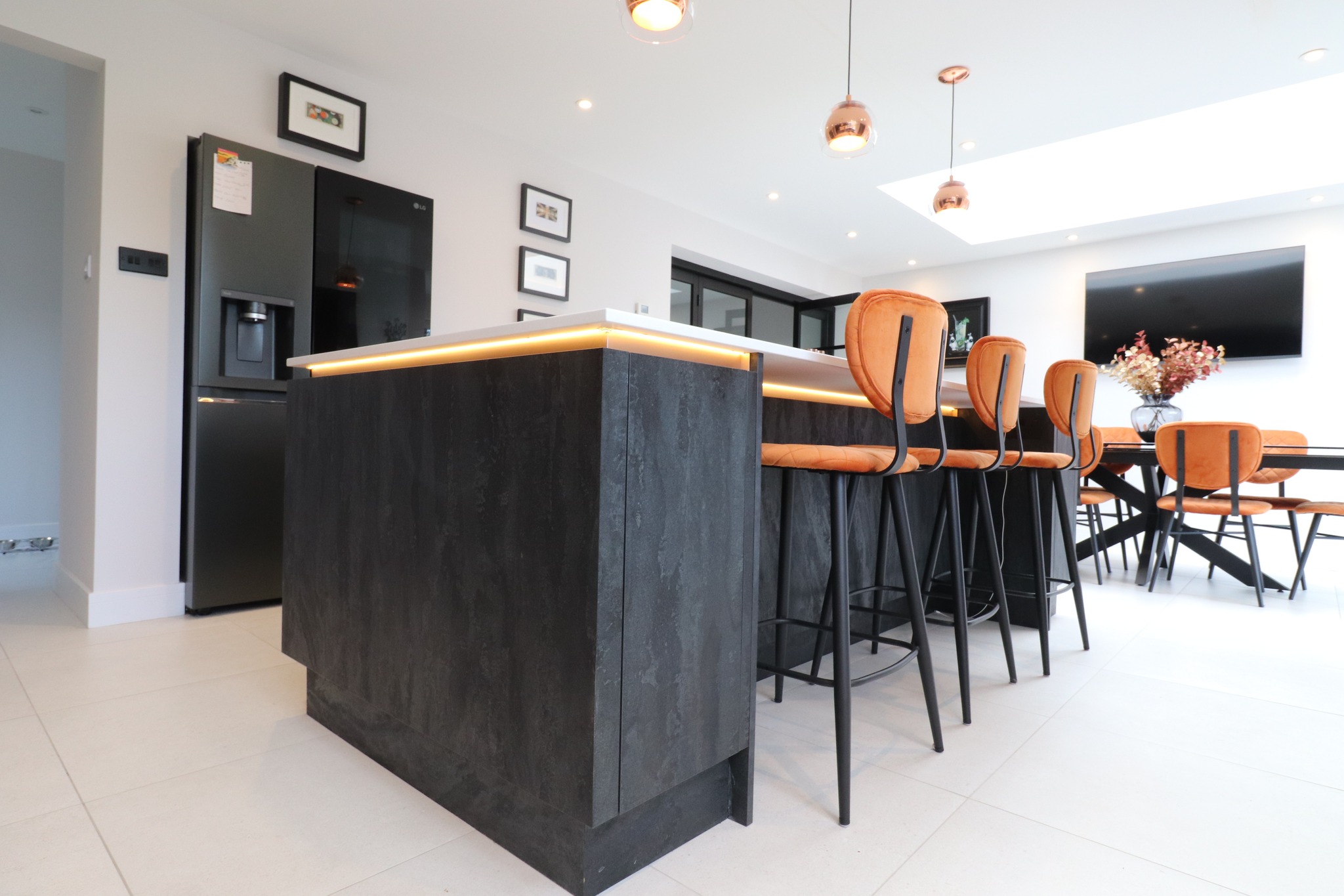 Dark textured handleless kitchen island with LED edge lighting, tan leather bar stools and copper pendants in open-plan kitchen diner — Inner Peace Interiors, Camberley