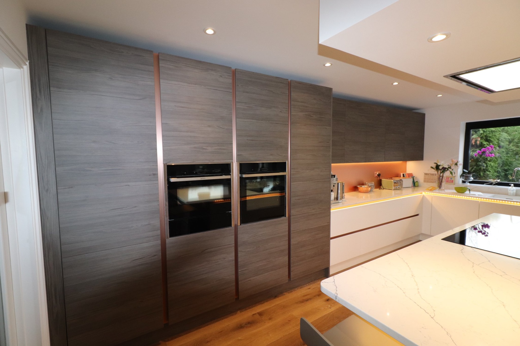 Dark wood handleless kitchen with integrated ovens, white worktops and warm under-cabinet lighting